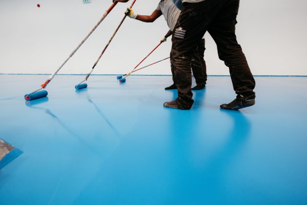 Construction workers painting a blue epoxy resin floor in a sports hall using rollers