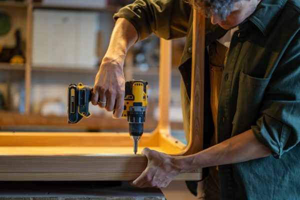 Carpenter attaches wooden table base to tabletop using power drill in joinery workshop. Handyman precision work, furniture assembly, custom design, professional woodwork techniques, handmade furniture